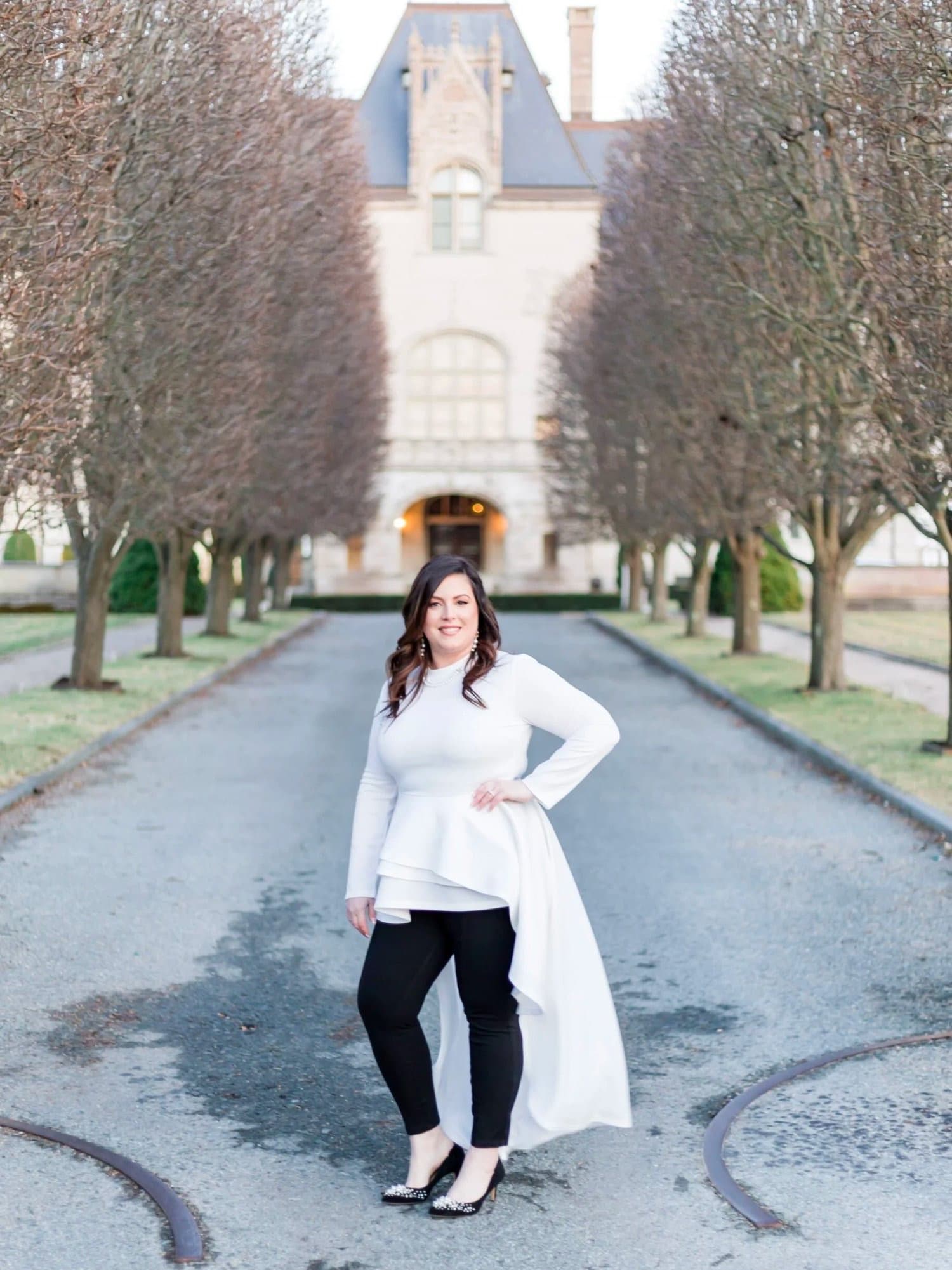Kate Daher standing on a tree-lined drive in a white top and black pants, smiling at the camera